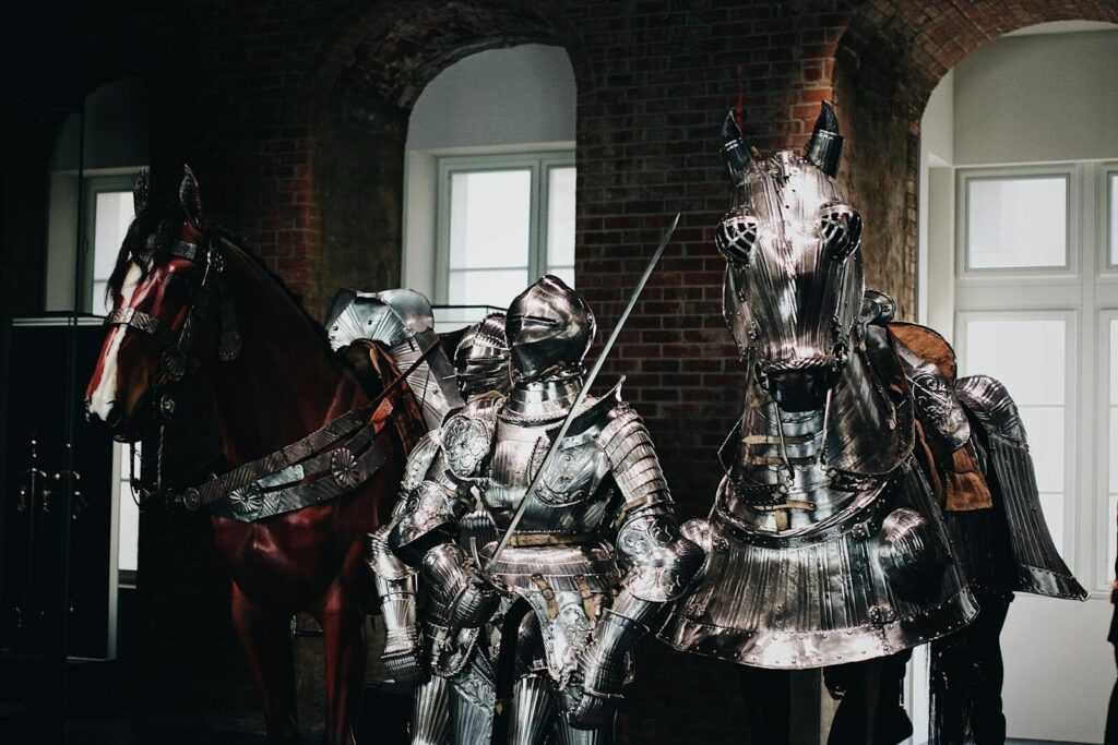 Medieval knight in full armor with a swordsman and armored horse indoors.