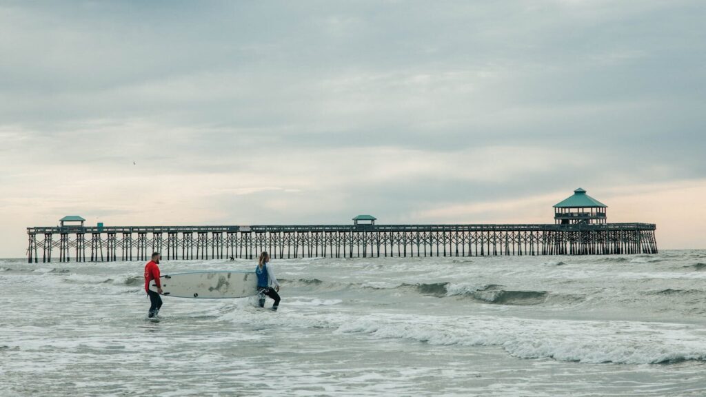 Two surfers carrying boards near Folly Beach Pier, SC, on an overcast day.