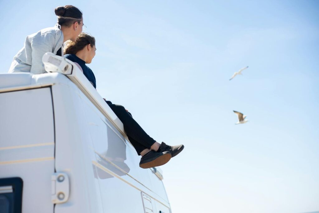 Two people sitting on a camper van roof enjoying a sunny day with seagulls flying.