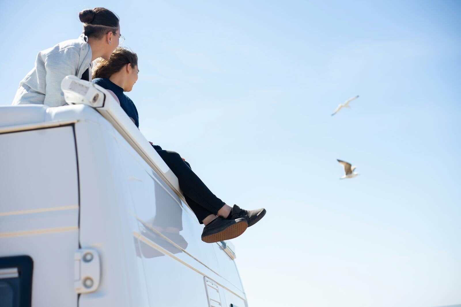 Photo by Kampus Production Two people sitting on a camper van roof enjoying a sunny day with seagulls flying.
