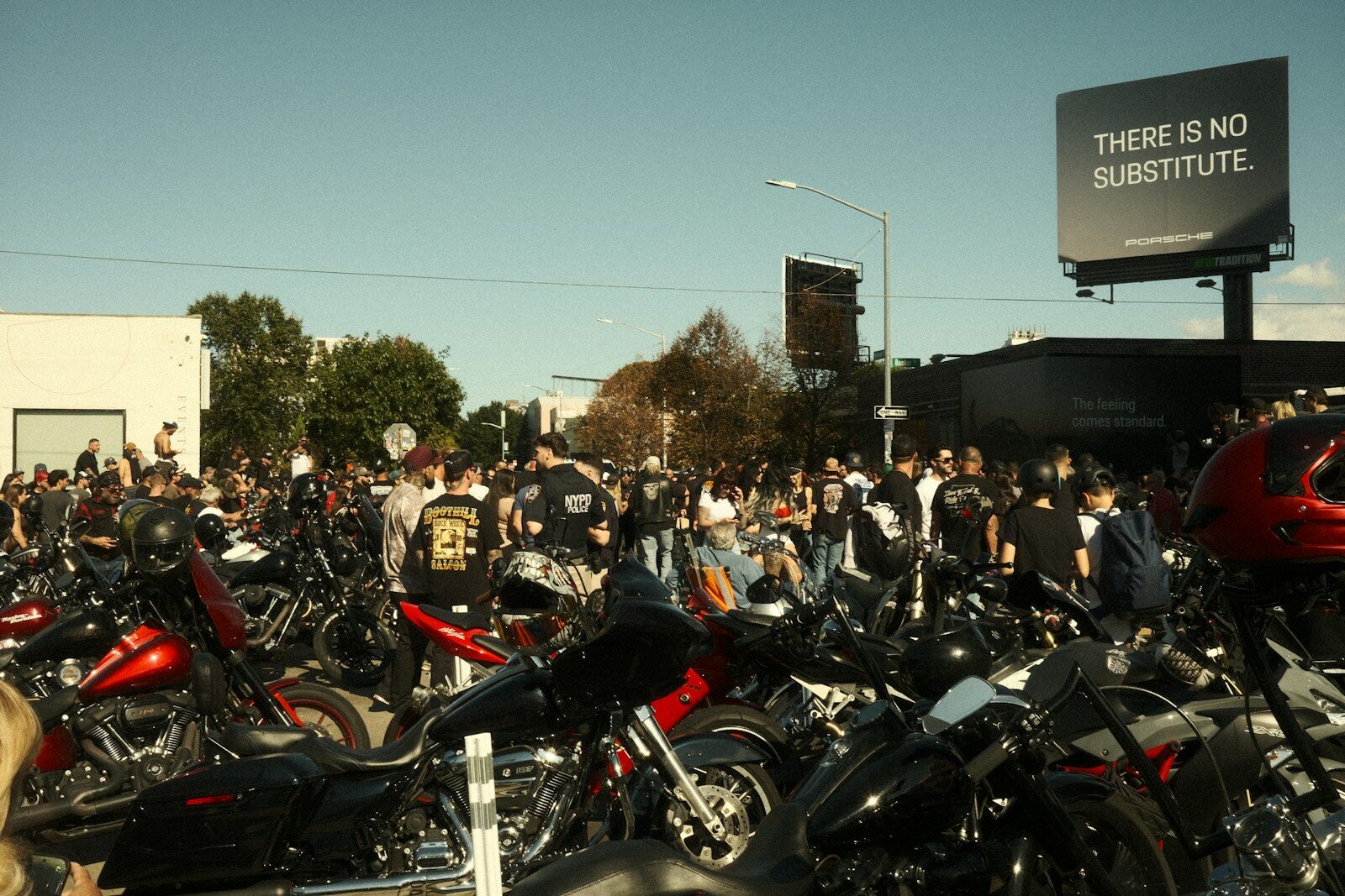 Crowd gathered around many motorcycles with a billboard.